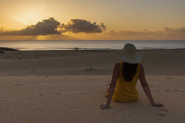 A woman is sitting on the beach wearing a straw hat. The sky is orange and the sun is setting