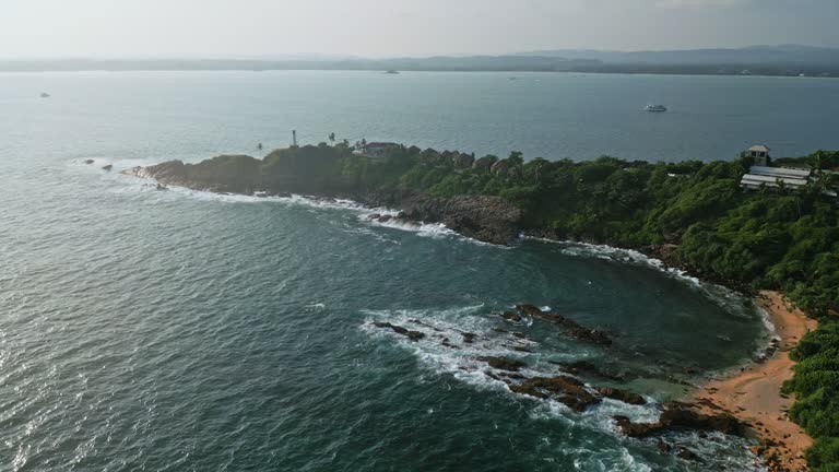 Drone view of coastal landscape with lighthouse on promontory, waves crash on rocky shore. Nautical beacon guides boats, ships at scenic bay. Tourists visit for adventure, lighthouse history, nature.