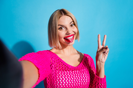 Photo portrait of lovely young lady selfie photo tongue out v-sign wear trendy pink knitted garment isolated on blue color background.