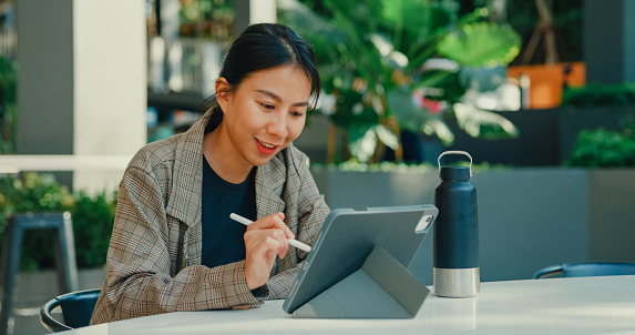 Young Asian businesswoman using a digital tablet for market research, sitting alone outdoors with a sustainable water bottle. Sustainable business outdoor concept.