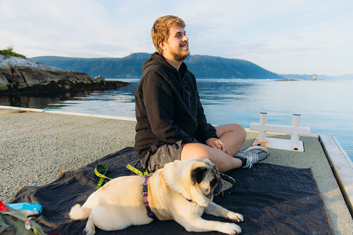 Side view of smiling man in black hoodie sitting on the pier with his pug enjoying summertime by the fjord with mountain view in Western Norway, Scandinavia