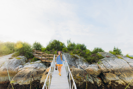 Front view of female in orange shorts walking down the wooden pier to the sea for picnic during summer sunset in Norway