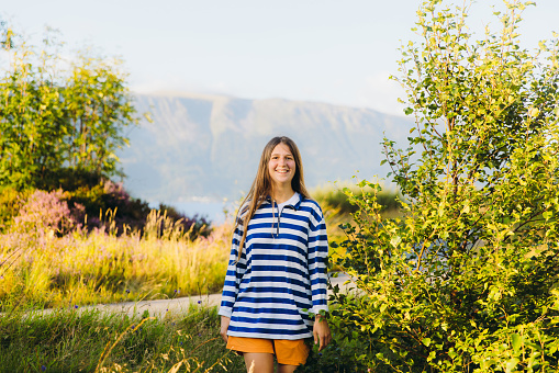 Portrait of smiling female traveler in striped shirt walking on meadow with  beautiful summer mountain landscape during sunset in Scandinavia
