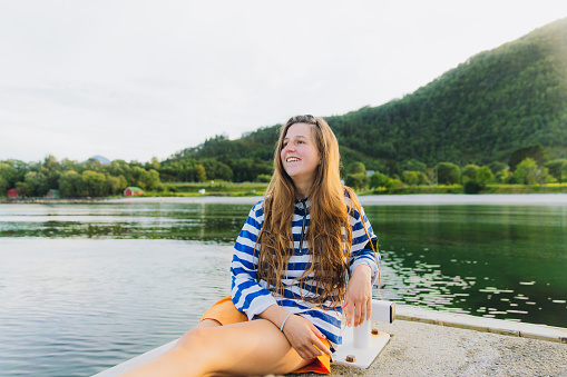 Smiling female sitting pn the pier with background view of scenic beach with green Mountain View during beautiful summer sunset