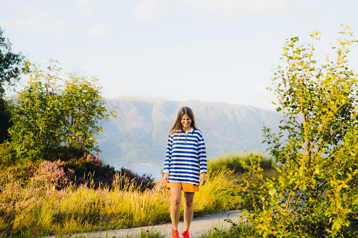 Portrait of smiling female traveler in striped shirt walking on meadow with  beautiful summer mountain landscape during sunset in Norway