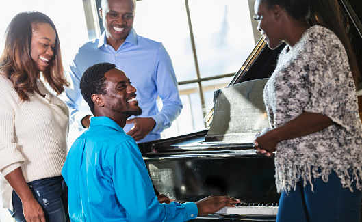A group of four African-American friends, two couples, hanging out together. One of the men is playing a piano, looking up at his partner. They are all smiling.