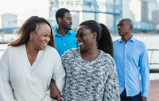 A group of four African-American friends, two couples, walking on a city waterfront, conversing and sightseeing. They are in their 30s and 40s. The focus is on the two women in the foreground, best friends walking arm in arm, smiling. Their partners are walking behind them, out of focus.