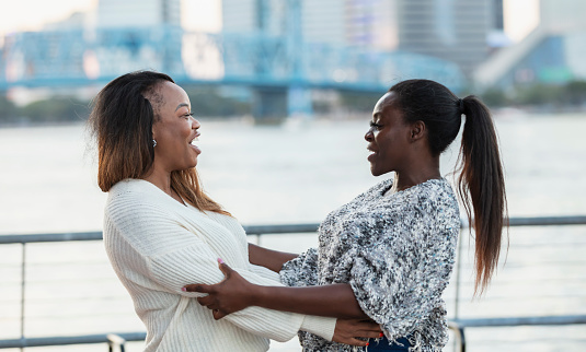 Two mature African-American woman, in their 40s, standing on a city waterfront, old friends greeting each other.