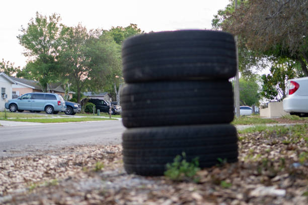 A dramatic close-up of a stack of four worn-out car tires abandoned by the roadside in a suburban neighborhood, illustrating themes of waste, environmental concern, and urban abandonment.