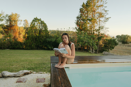 Photo of a young woman reading a book by the pool