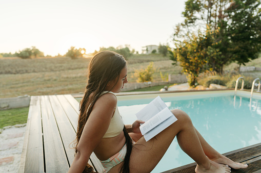 Photo of a young woman reading a book by the pool