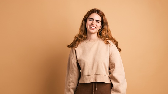 Portrait of cheerful young woman in trendy clothes with long dark hair smiling at camera while standing against brown background