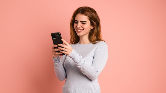 Smiling young woman in casual clothes with dark hair text messaging on mobile phone while standing against pink background