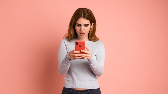Shocked young woman in casual clothes with dark hair using smartphone while standing on pink background