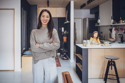 Portrait of a beautiful Japanese woman standing at home, her daughter is sitting in kitchen behind her.