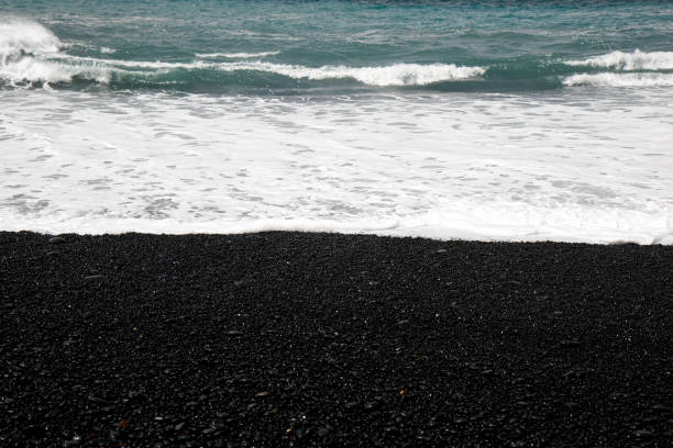 Black volcanic sand beach displaying smooth contrast between wet and dry surfaces with scattered seashell fragments. Ocean waves shaping organic shoreline patterns with rich natural textures. Captured in Taiwan, Asia.