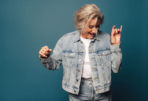 Happy, mature woman in casual denim stands in a studio, smiling and having fun while dancing. Fashionable older woman moving with grace and embodying a young-at-heart lifestyle.