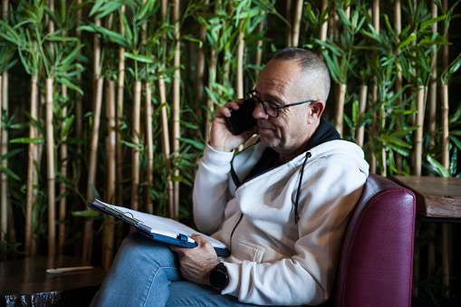 Side View Of Young Man Filling Out Form While Sitting With Plants Behind Him Talking On The Phone