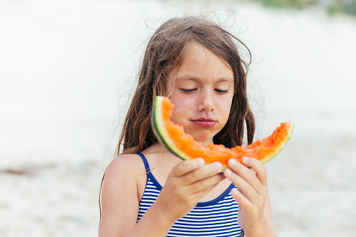 Child holding slice of watermelon and eating as a snack on her summer vacation at the beach