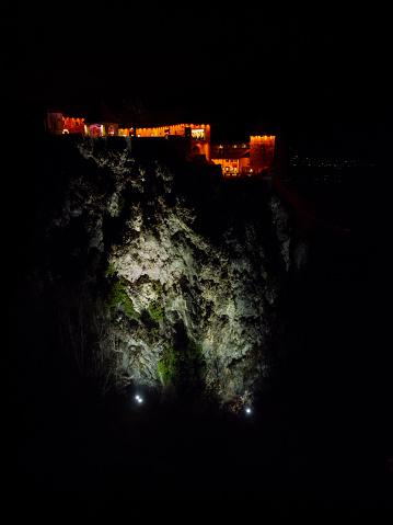 Scenic and dramatic view of illuminated Lake Bled Castle on top of the rock at night. Front view of lights on the rock and orange lights showing silhouette of the castle. Date: 19.12.2023, Bled, Slovenia.