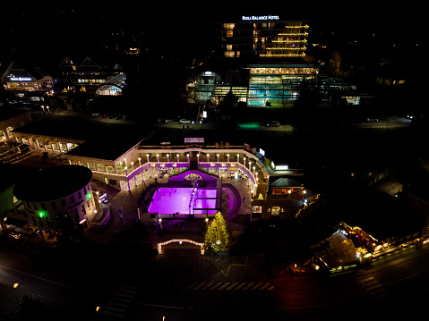 Bled town ice skating with purple lights at Christmas time. Aerial view above ice skating ring within Bled town shopping center. Hotels with indoor swimming pools behind. Date: 19.12.2023, Bled, Slovenia.