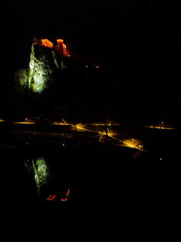 Illuminated Lake Bled Castle and the rock at night with clear reflection on the water surface. Walkway with street lights. Date: 19.12.2023, Bled, Slovenia.