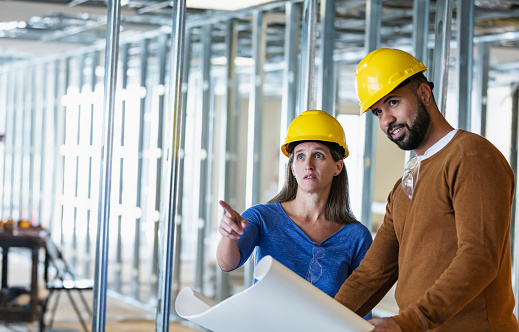 Two workers wearing hardhats conversing at a construction site, an office space being renovated for a new tenant. The African-American man, in his 30s, is holding open a set of blueprints. His coworker, a mature woman in her 40s, is looking up, pointing at something. The main focus is on the woman.