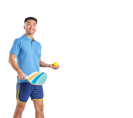 asian boy in shorts and blue polo shirt with a paddle and pickleball ball in his hands on white background