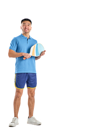 smiling asian young man in sports shorts and blue polo shirt holding pickleball paddle in hands on white background