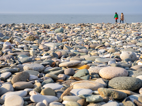 Gray sand and stones on the beach. Two silhouettes of people on the shore in the distance. Beach in winter. Sea coast soil. Beach resort atmosphere