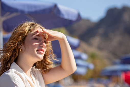A woman with blonde hair is looking up at the sky. She is wearing a white shirt and a gold necklace. The scene is set on a beach with blue umbrellas and palm trees in the background