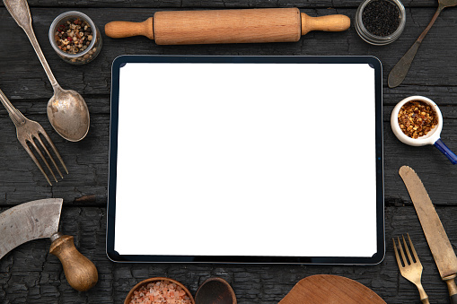 Tablet with blank screen and kitchen and cooking utensils on black wooden table.