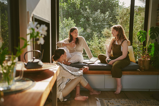 Smiling group of diverse female friends talking while sitting together at a bench by a window during a women's wellness retreat