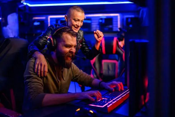 Young bearded male cybersport gamer playing video game in a gameroom and celebrating victory with his female friend. Esport streaming game online. Bald woman is hugging him and cheering. Copy space.