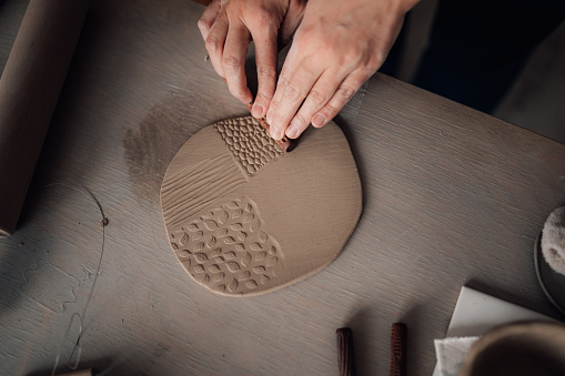 Cropped picture of an unrecognizable craftswoman using decorative rolling pin for clay work and clay decorating. Close up of craftswoman's hands imprinting pattern and decorating clay on pottery class