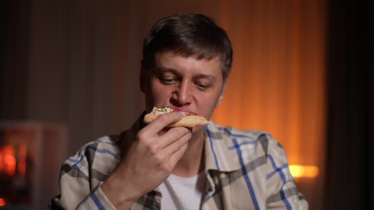 Hungry handsome young man biting delicious slice of pizza sitting alone at table in evening or night on blurred background of soft light.