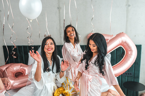 In the hotel room, diverse bridesmaids and a radiant Hispanic bride, all in silk bathrobes, share playful moments with balloons during bachelorette party preparations.