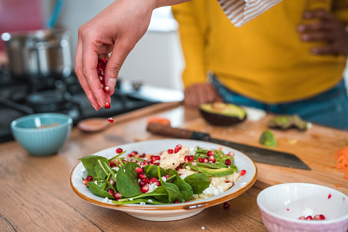 An unrecognizable woman puts together a colorful salad plate in a close-up photo. With fresh spinach, grated carrots, chickpeas, avocado, feta cheese, and pomegranate seeds she transforms a salad into a food masterpiece.