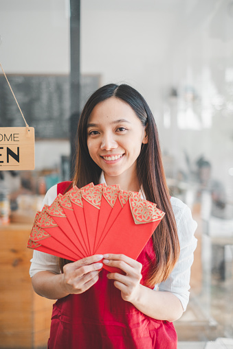 A bright-eyed young woman in an apron presents traditional red envelopes, her smile reflecting the cultural joy and hospitality associated with this gesture.