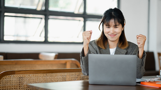 Joyful female professional triumphantly raises her fists in victory while working on a laptop in a bright office.
