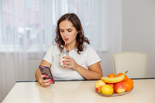 Young woman is browsing her smartphone while enjoying a banana mascarpone smoothie at home with fresh fruits on the table.