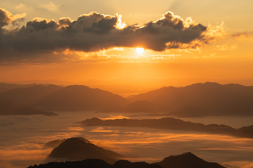 The sun is setting over a mountain range, casting a warm glow over the landscape. The sky is filled with clouds, creating a sense of mystery and depth. The mountains are covered in fog