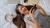 top view of asian woman lying on bed wearing headphone enjoy playing online mobile games at home