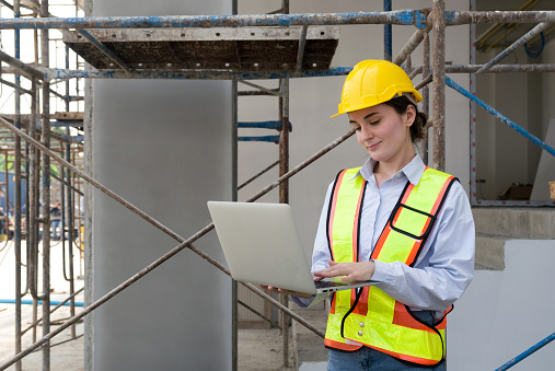 A worker with a helmet stands by metal scaffolding, typing on laptop computer.