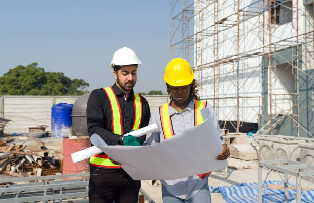 istock Two construction worker looking at a floor plan, standing in front of a large building site with helmet and safety gear. Day time work safety check. Work environment at the site of housing projects. 2098110961