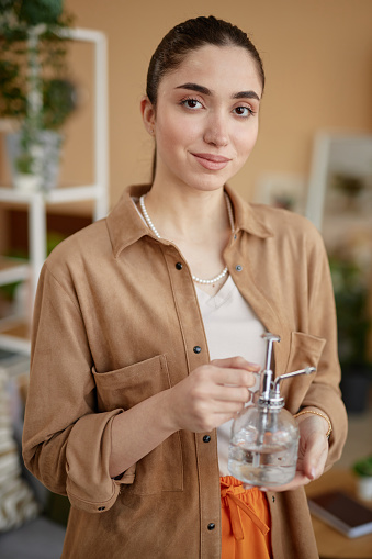 Vertical portrait of smiling woman holding glass watering can and looking at camera in cozy home