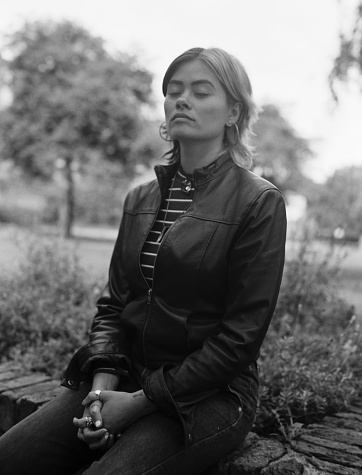 Nostalgic monochrome image of relaxed mixed race woman sitting on a bench in a park. Her eyes are closed and she is wearing leather jacket and jeans. Image taken with film analog camera.