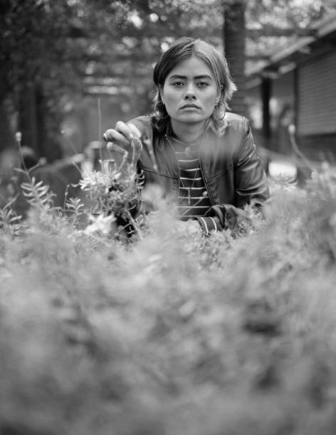 Intense mixed race woman looking at camera in a monochrome portrait. There are blurry vegetation in the foreground and a blurry building in the background. Medium format analog film camera.