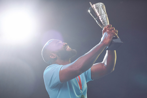 Side view portrait of African American sportsman holding trophy cup and cheering while celebrating victory with lens flare copy space