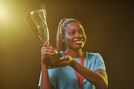 Waist up portrait of female African American athlete holding trophy cup and celebrating victory smiling happily
